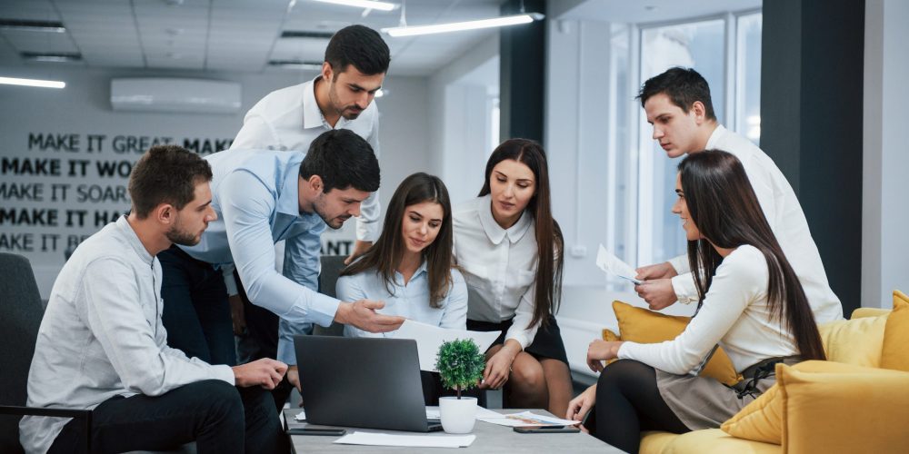 Guy shows document to a girl. Group of young freelancers in the office have conversation and working.